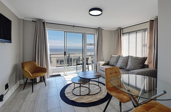 A modern living room with a sofa, two armchairs, a glass coffee table, and a view of the sea through large windows.