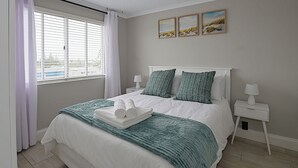 Une chambre à coucher avec un lit, des tables de chevet blanches, un ventilateur de plafond et une fenêtre avec des rideaux.