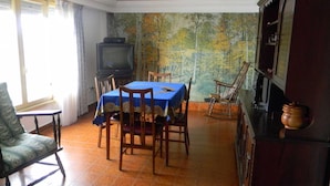 Ein Essbereich mit einem Tisch für vier Personen, einem Fernseher auf einem Standfuß und einer rustikalen Wand mit einem Waldgemälde.