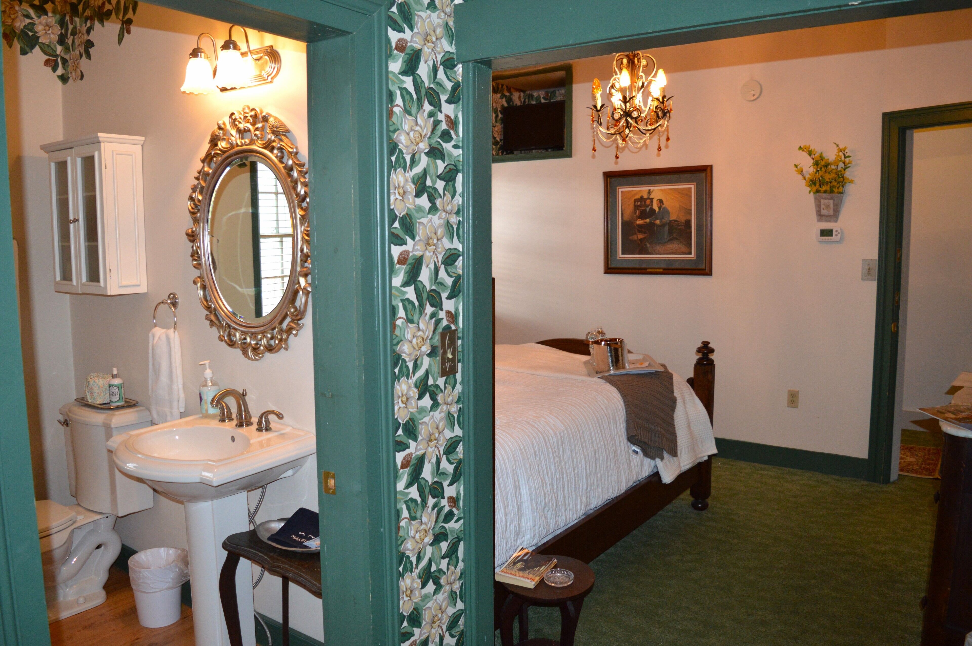 A bedroom with a bed, a bedside table, a nightstand, a dresser, a mirror, a chandelier, and a window.