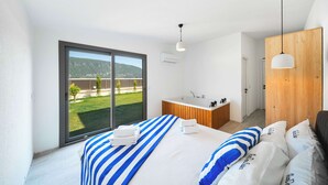 Ein Schlafzimmer mit einem großen Bett, einem Holzschrank, einem Badezimmer mit einer Badewanne und Blick auf eine grüne Landschaft durch eine Schiebetür.