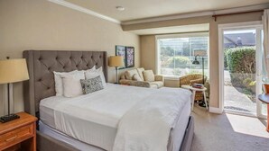 A bedroom with a large bed, a nightstand, a chair, a small table, and a window with blinds. - The Courtyard - Grand Queen Room at Cannon Beach Hotel Collection! Kitchenette (Cannon Beach)