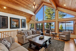 Amplia sala de estar con techo alto, ventanas grandes que ofrecen vistas a un lago y un bosque, y una terraza de madera en el exterior.