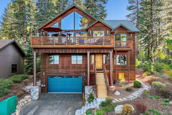 A two-story log cabin with a blue garage door, a balcony with outdoor seating, and a stone pathway leading to the entrance.