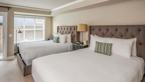 A hotel room with two beds, a nightstand, a lamp, and a window with blinds. - The Courtyard - Double Queen Room with Beach Access and Kitchenette! (Cannon Beach)