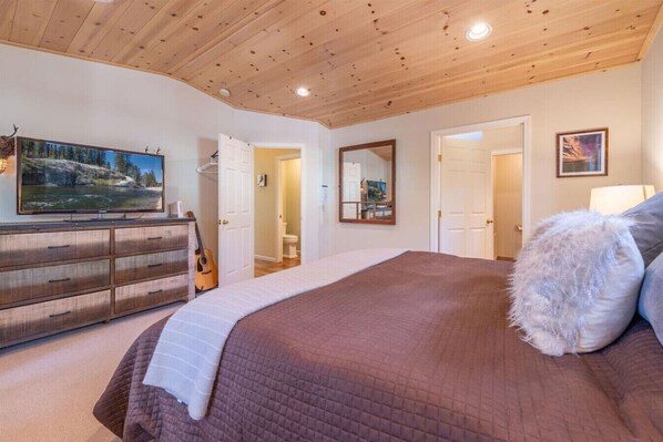 Un dormitorio con techo de madera, una cama grande con cubrecama acolchado, un aparador con una guitarra, un cuadro en la pared y un espejo.