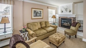 Un salón con un sofá color beige, un sillón a juego, un pequeño otoman, una chimenea y un cuadro en la pared.