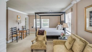 Un dormitorio con una cama con dosel, una zona de estar con un sofá y sillones, una mesita con una lámpara y una ventana con persianas.