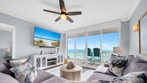 A living room with a large TV, a ceiling fan, and a view of the ocean through large windows.