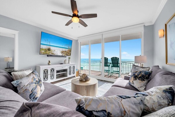 A living room with a large TV, a ceiling fan, and a view of the ocean through large windows.