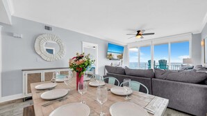 A dining area with a table set for six, a gray sofa, a ceiling fan, and a large window with a view.
