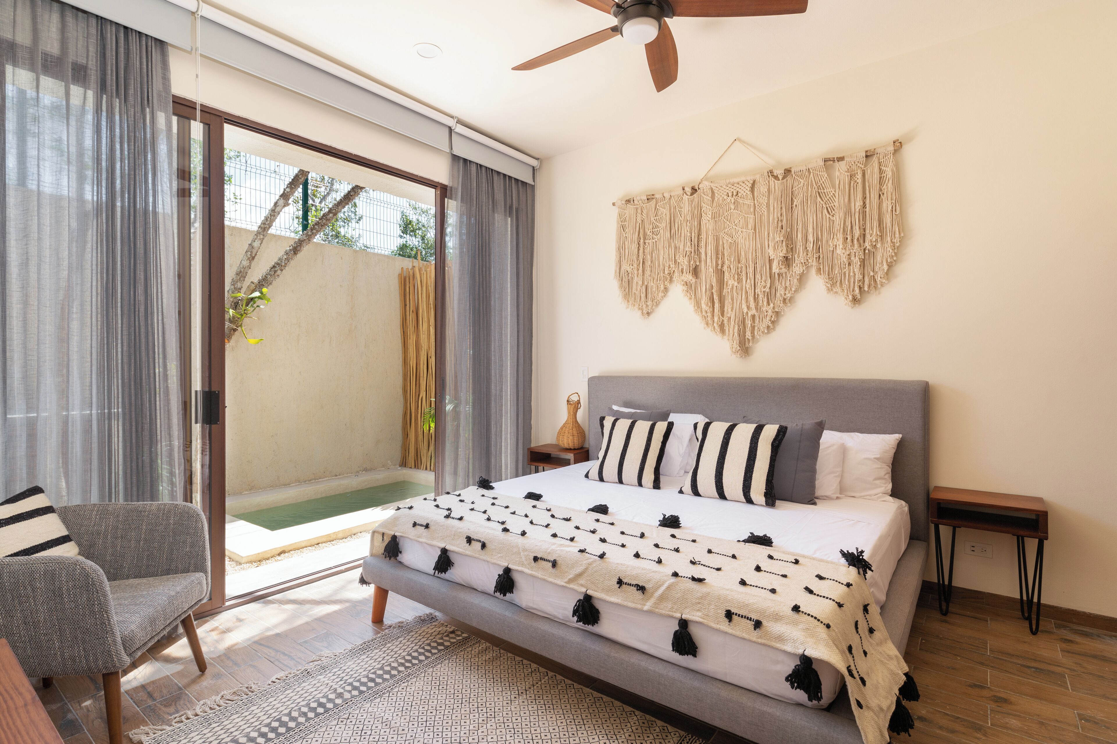 Un dormitorio con una cama, una silla, un ventilador de techo, un tapiz de macramé y vistas por un cristalero corredizo.