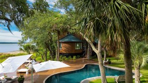 A wooden house with a pool, surrounded by lush greenery and a view of the water.