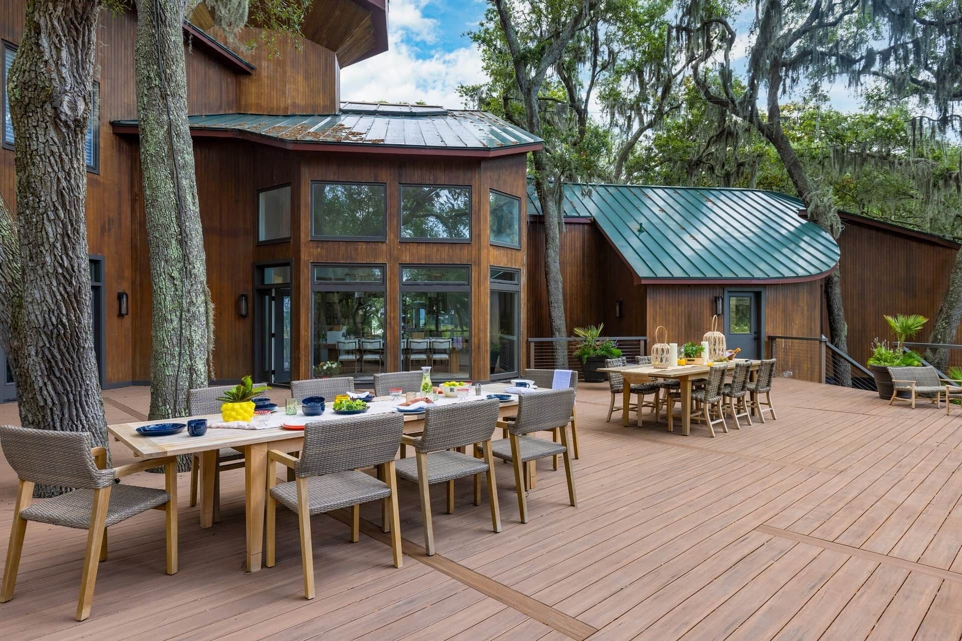 A wooden deck with a dining area, surrounded by trees and a building with large windows and a green roof.