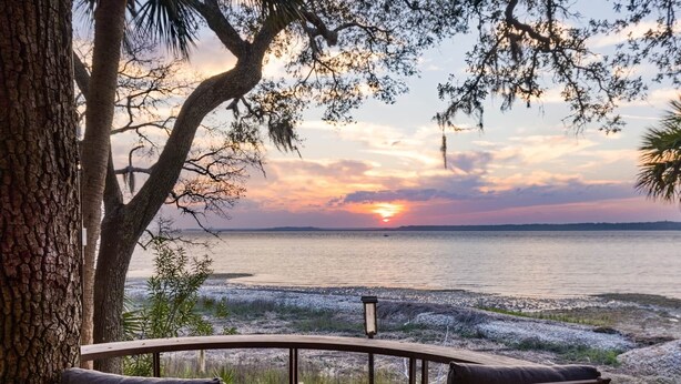A wooden deck with two chairs and a table, overlooking a body of water with a sunset in the background.