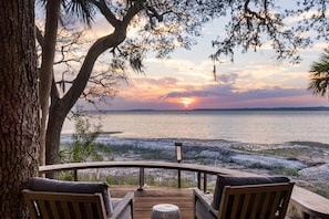 A wooden deck with two chairs and a table, overlooking a body of water with a sunset in the background.
