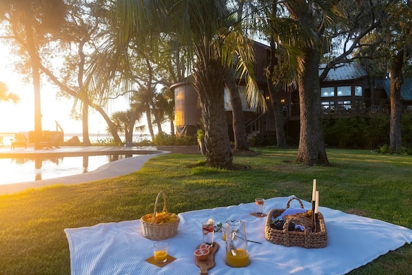 A picnic setup with a table, baskets, and drinks on a grassy area near a pool and palm trees.