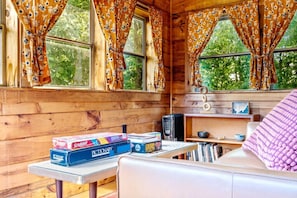 Interior acolhedor de uma cabana, com paredes de madeira, uma mesa com jogos de tabuleiro, uma cafeteira e uma janela com cortinas.