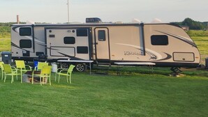 A Kodiak RV with a flatbed trailer, a table, and four chairs in a grassy field.