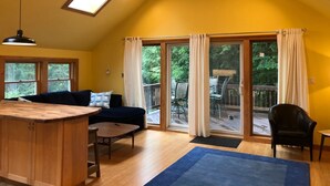 Un salon chaleureux avec un plafond en bois, un ventilateur de plafond, une grande fenêtre avec des rideaux, un canapé bleu foncé, une table basse en bois et une petite chaise noire.