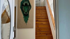 Un couloir avec un escalier en bois, une porte dotée d’une fenêtre ronde et une œuvre d’art représentant une créature verte, accrochée au mur.