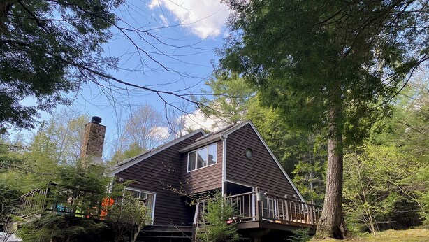 Une cabane avec une cheminée en pierre, une terrasse et entourée d’arbres.