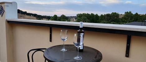 A table with a bottle of wine and two glasses on a balcony with a view of trees and buildings.