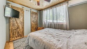 Un dormitorio con una cama grande, ventilador de techo, televisión montada y ventana con cortinas.