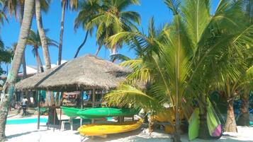 Escena de playa con una cabaña de techo de paja, palmeras y varios kayaks y canoas de colores.