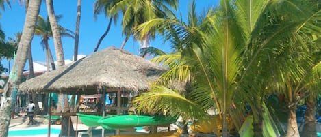 A beach scene with a thatched-roof hut, palm trees, and several colorful kayaks and canoes.