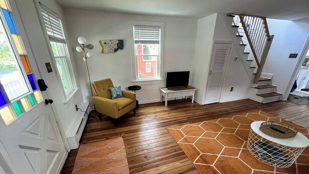 A living room with wooden flooring, a yellow armchair, a TV, and a staircase. - Charming 2-BRM Cottage in Tranquil Island Heights - Steps from the River (Island Heights)