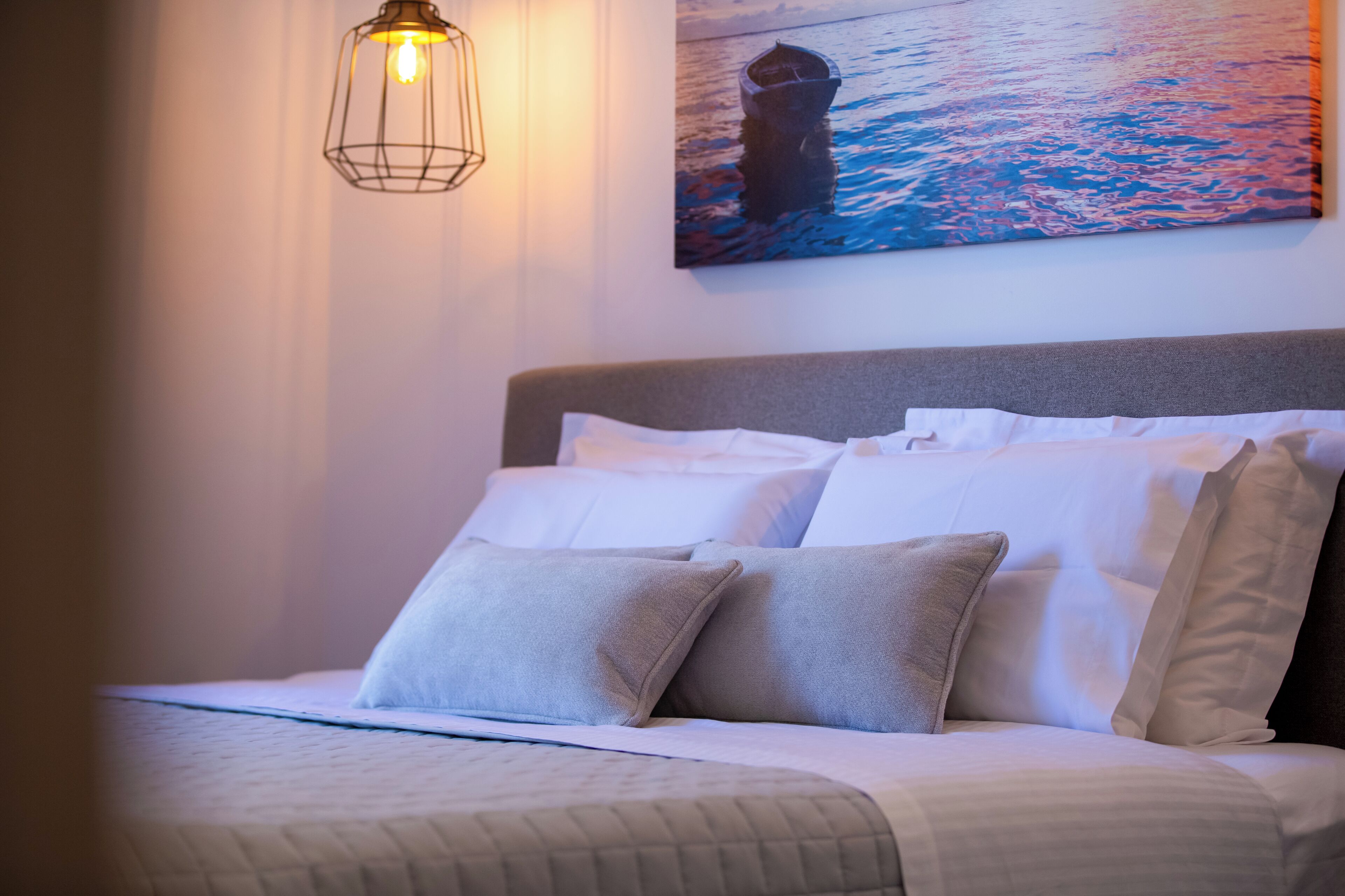 A neatly made bed with white and grey bedding, a wall-mounted light fixture, and a painting of a boat on water.