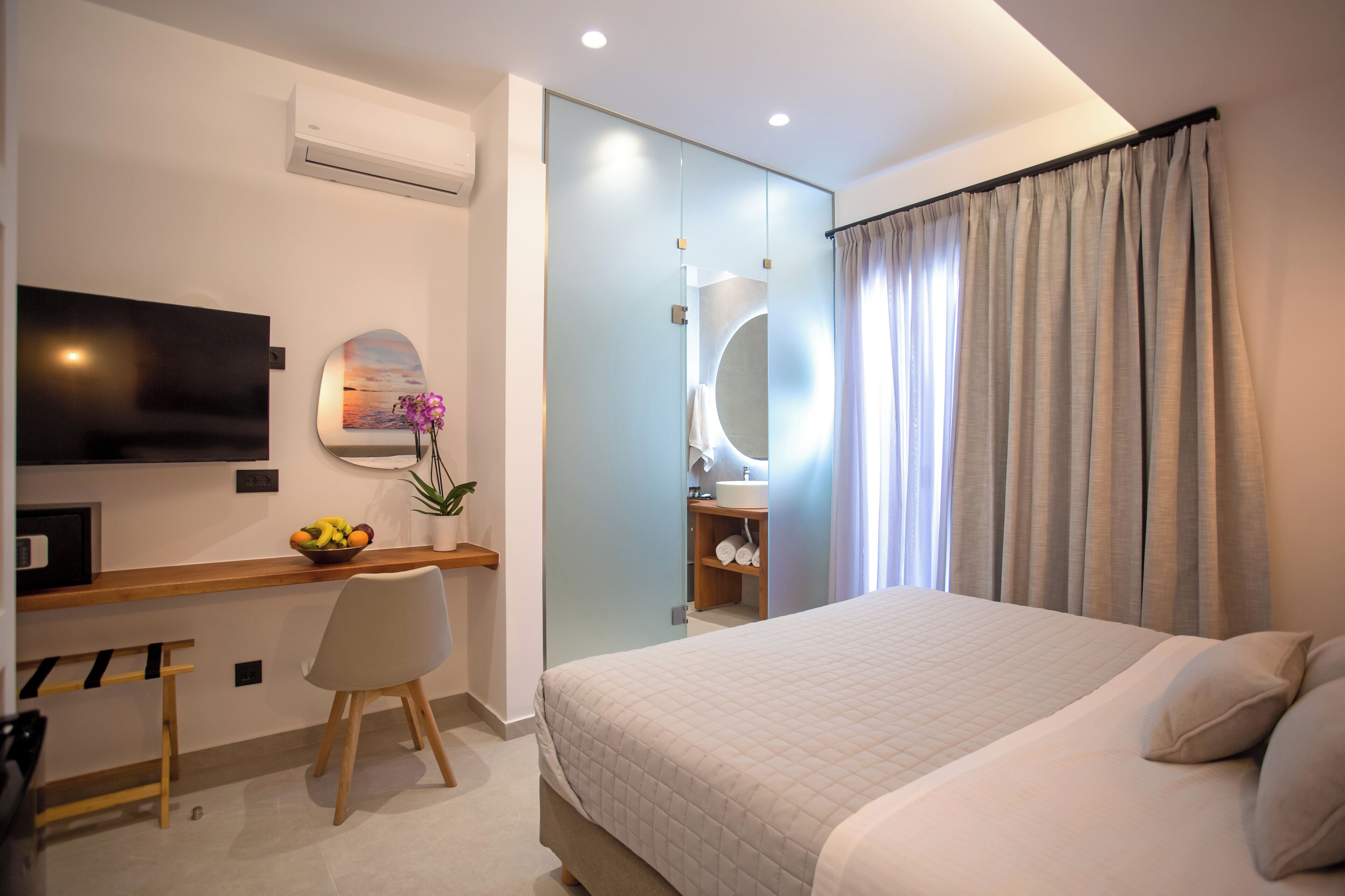 A modern hotel room with a large bed, a desk with a chair, a TV, a mirror, and a bathroom visible through a glass partition.