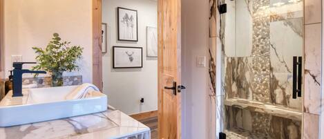 A bathroom with a marble sink, a potted plant, framed artwork, and a glass-enclosed shower with a marble design.