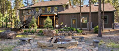A two-story lodge with a stone fire pit, outdoor seating, and a staircase leading to a deck.