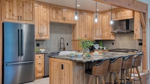 A modern kitchen with wooden cabinets, a stainless steel refrigerator, a marble island, and bar stools. - Spring River Retreat by Avantstay Lakefront Luxury w/ Hot Tub, Game Room & Near Bend (Bend)