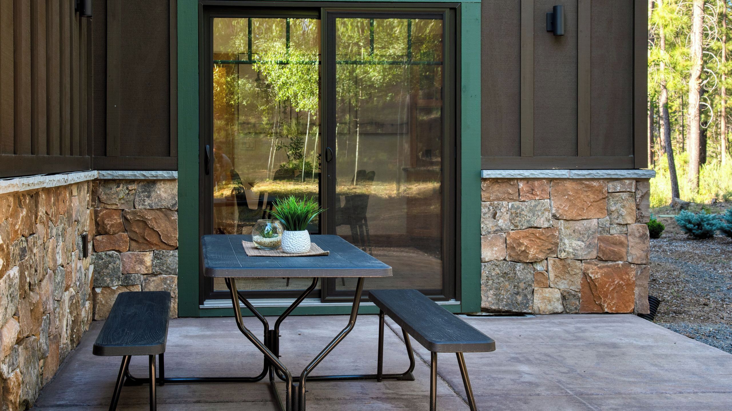 A patio with a table and benches, a stone wall, and a glass door leading to a garden.