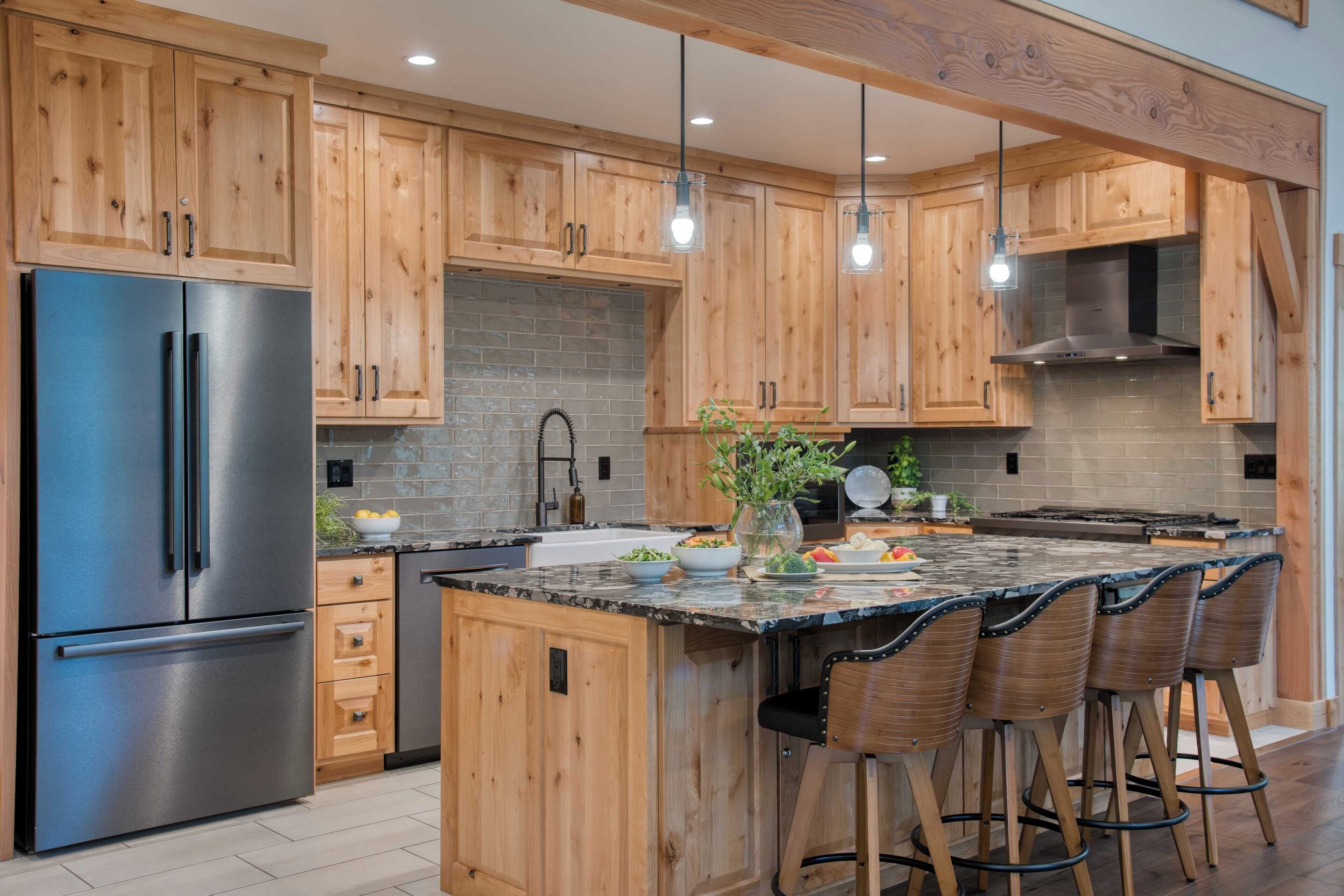 A modern kitchen with wooden cabinets, a stainless steel refrigerator, a large island with bar stools, and pendant lighting.