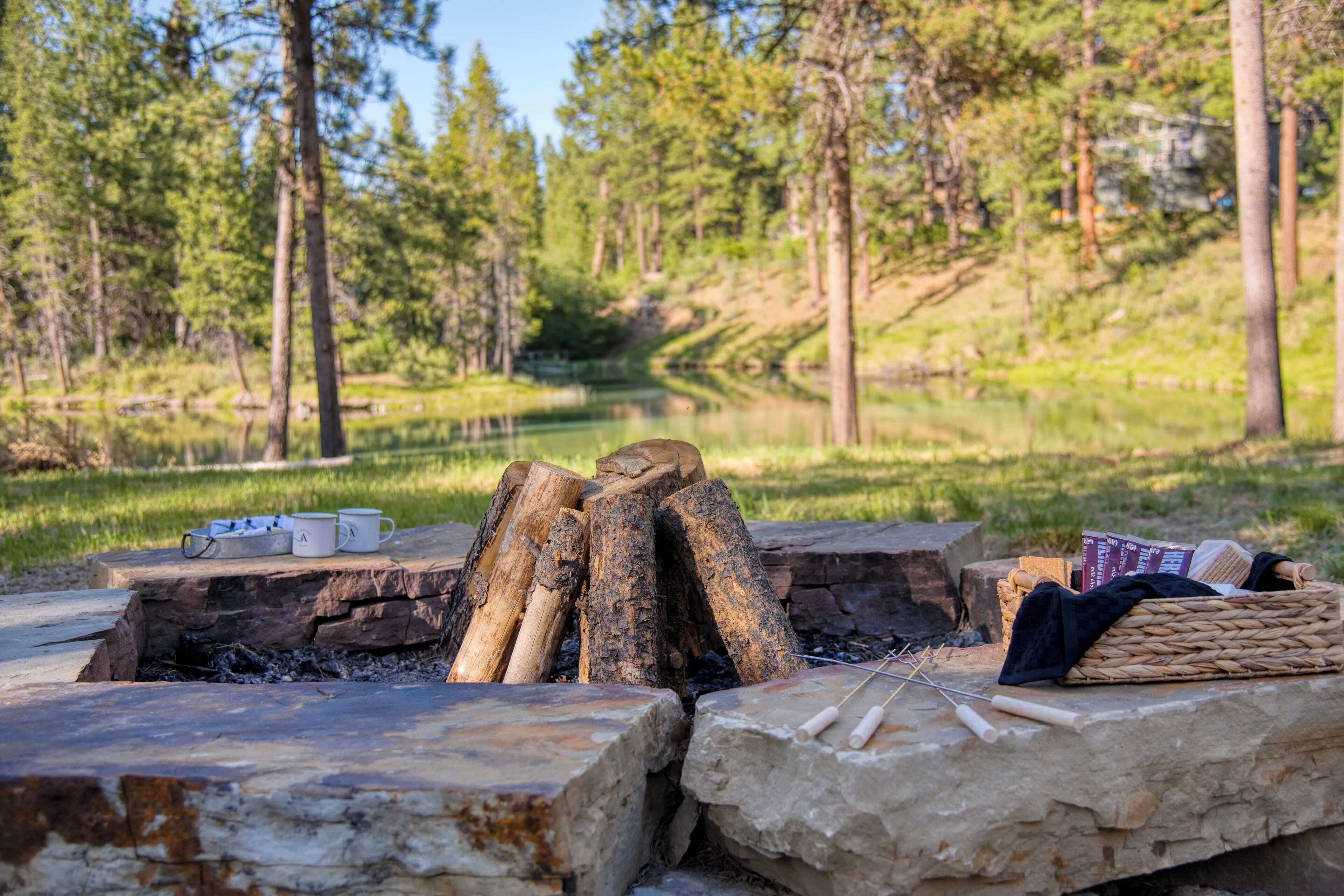 A campfire setup with logs, stones, and camping gear in a forested area with a river in the background.