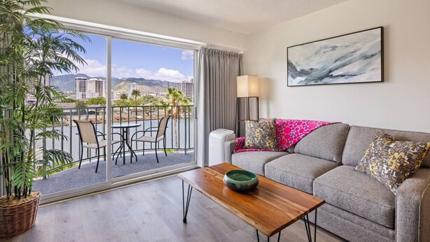 Una sala de estar moderna con un sofá gris, una mesa de centro de madera y vistas a un balcón con mobiliario exterior y un paisaje urbano.