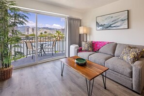A modern living room with a grey sofa, a wooden coffee table, and a view of a balcony with outdoor seating and a cityscape. - 401 FREE Parking/10 min beach/2b-Waikiki/Ala Moana (Honolulu)
