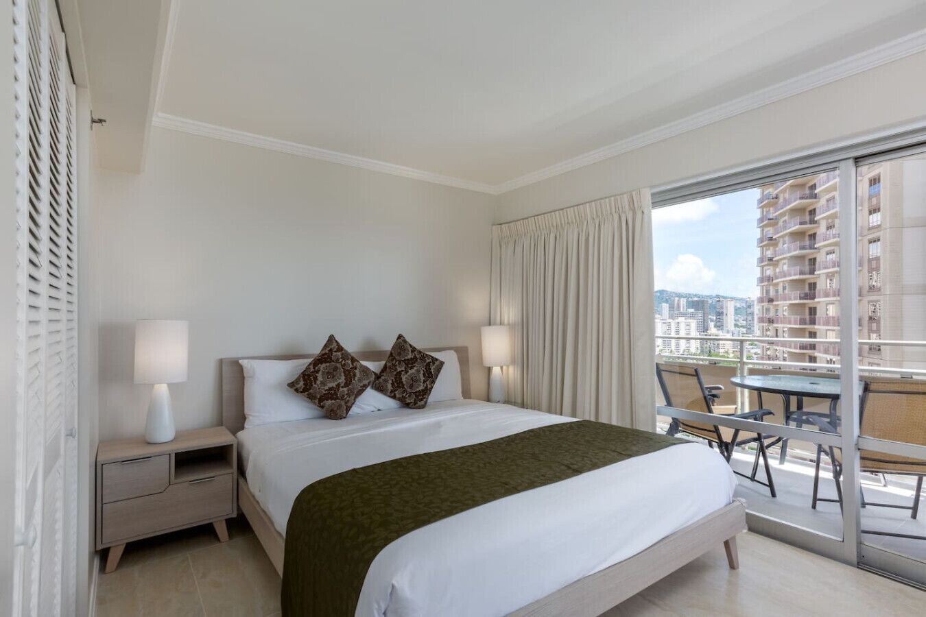 Una habitación de hotel con una cama grande, dos lámparas de noche, una mesita de noche, un plaid verde y un balcón con mesa y sillas.