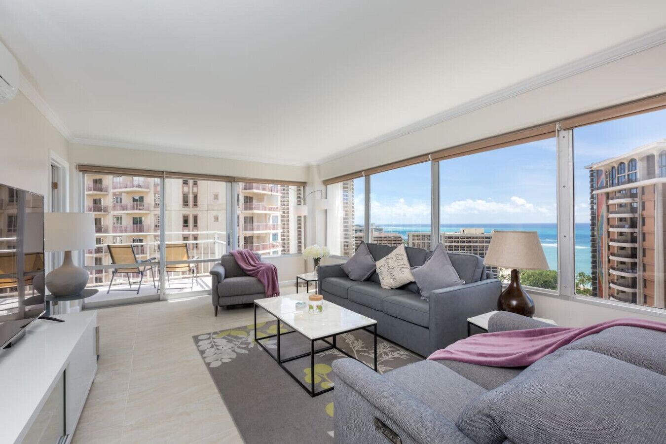 A spacious living room with a large window, a gray sofa, a coffee table, and a view of the ocean and buildings.