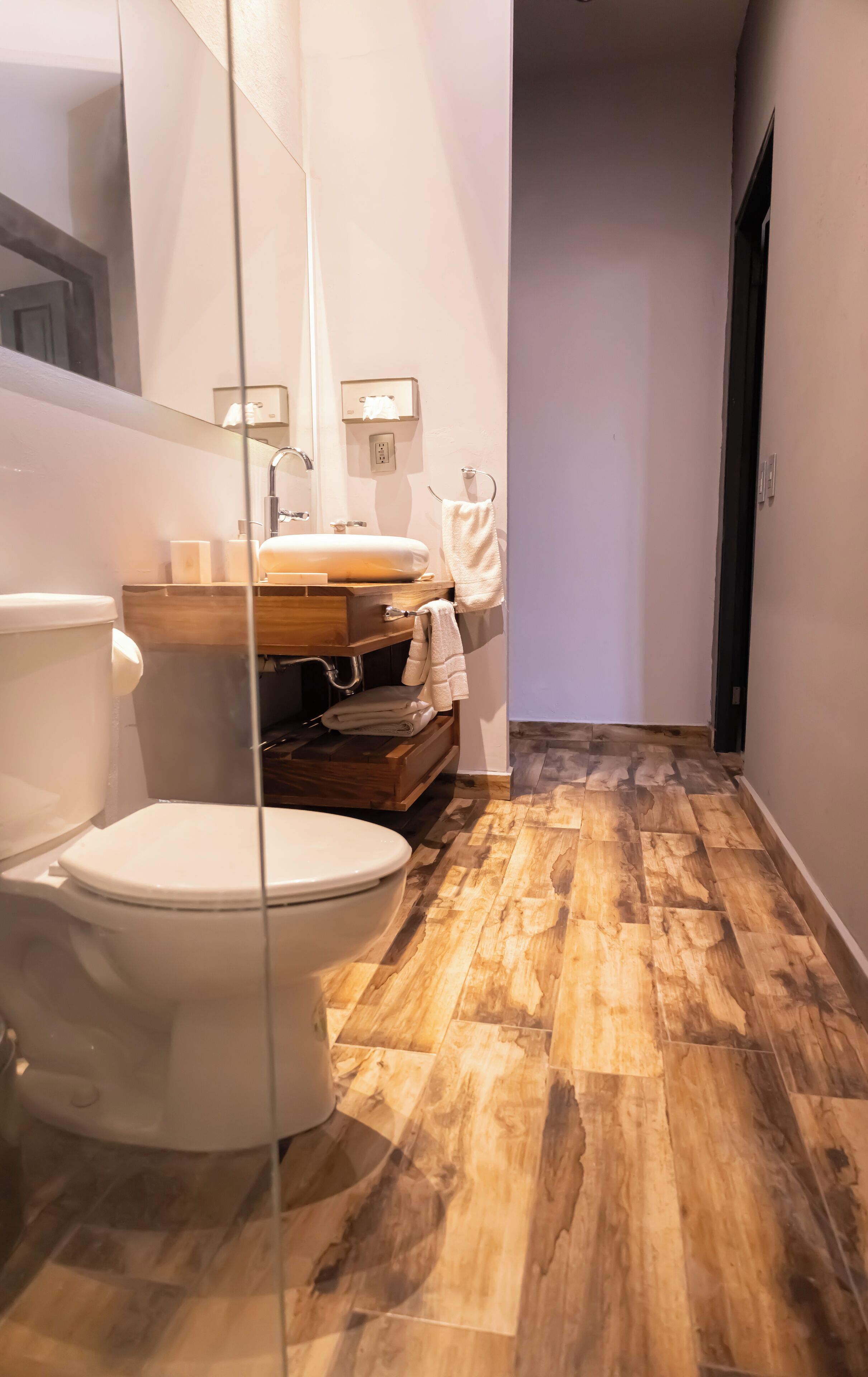 Un baño moderno con inodoro, lavamanos y espejo. El suelo tiene un diseño que imita la madera.