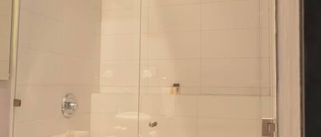 Un área de ducha con paredes de vidrio, un muro revestido de azulejos blancos y un cabezal de ducha visible, con un inodoro a la izquierda.