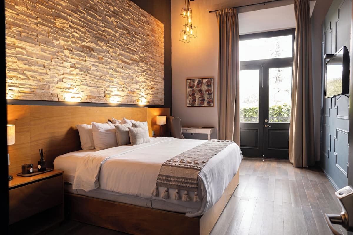 Un dormitorio moderno con pared de piedra, una cama grande, mesitas de noche y vistas a través de una puerta.