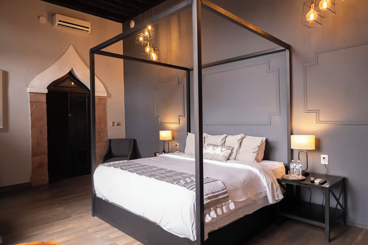 Una cama con dosel, ropa de cama blanca y gris, dos mesitas de noche con lámparas, una pared de piedra y una puerta.