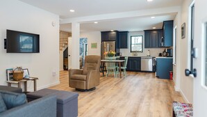 A modern kitchen with dark cabinets, a dining area with a table and chairs, and a living room with a sofa and a TV mounted on the wall. - Downtown Freeport Barn'acle (Freeport)