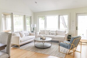 A spacious living room with a large window, a round coffee table, and a grey armchair.