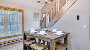 Un coin repas avec une table en bois dressée pour quatre personnes, un lustre et une vue sur l’extérieur grâce à de grandes fenêtres.